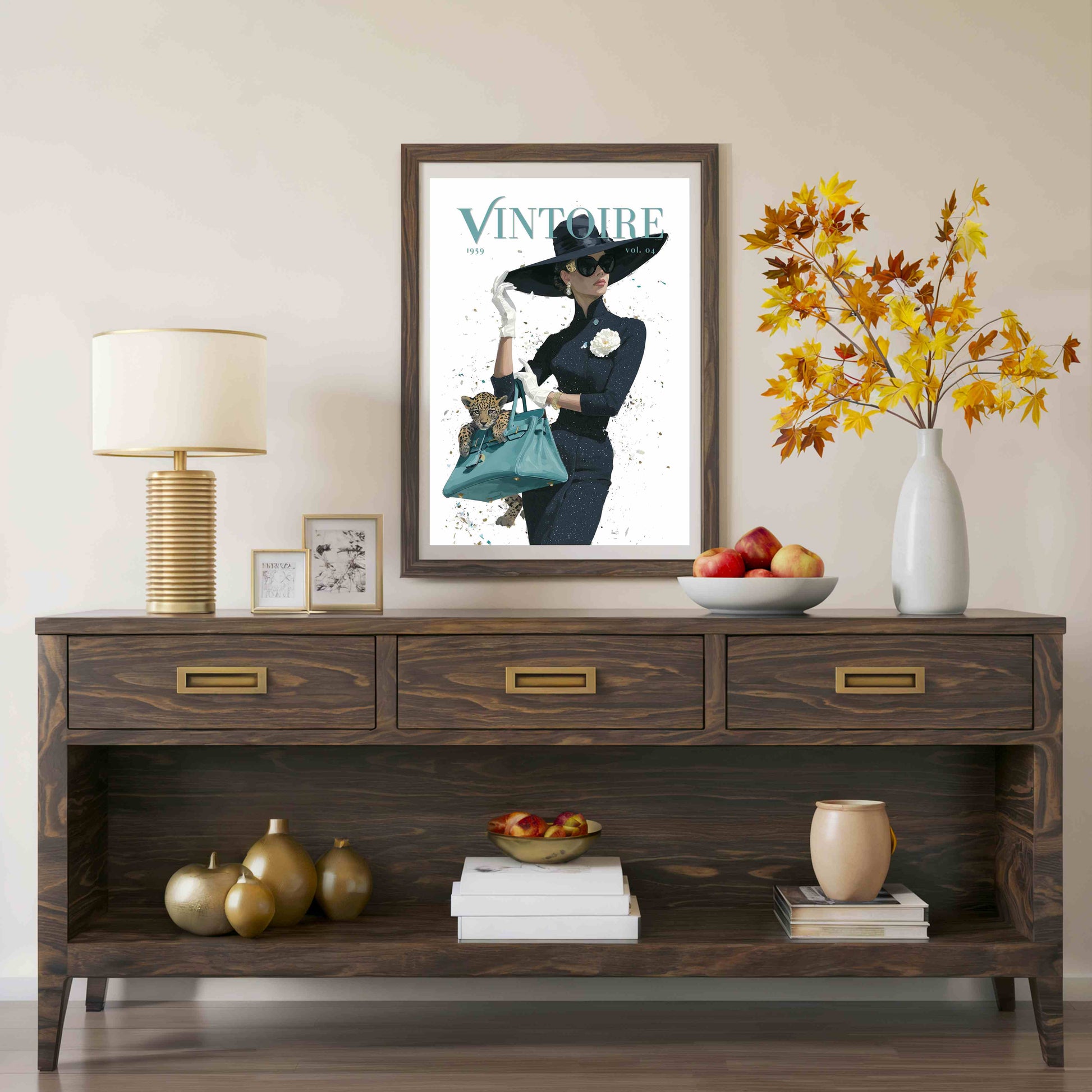 A framed vintage-style poster with the word 'VINTOURE' hangs above a dark wood console table in a styled interior. The room includes a table lamp, small framed art, a bowl of fruit, a vase with autumn leaves, and decorative objects on the table and shelf.