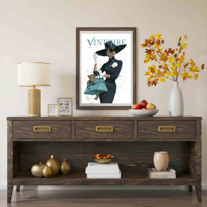A framed vintage-style poster with the word 'VINTOURE' hangs above a dark wood console table in a styled interior. The room includes a table lamp, small framed art, a bowl of fruit, a vase with autumn leaves, and decorative objects on the table and shelf.