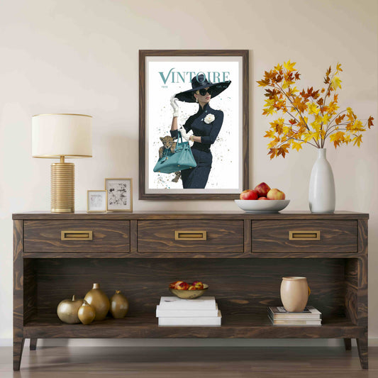 A framed vintage-style poster with the word 'VINTOURE' hangs above a dark wood console table in a styled interior. The room includes a table lamp, small framed art, a bowl of fruit, a vase with autumn leaves, and decorative objects on the table and shelf.