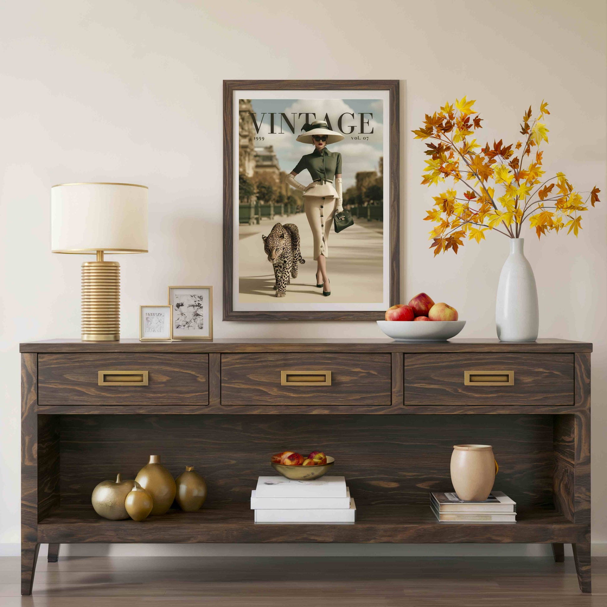 A dark wood console table styled in a living room setting, featuring three drawers with brass handles and an open lower shelf. The table is decorated with a table lamp, picture frames, a bowl of apples, a vase with autumn leaves, and decorative gourds.