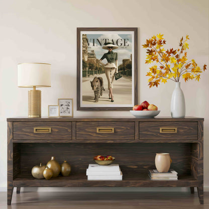 A dark wood console table styled in a living room setting, featuring three drawers with brass handles and an open lower shelf. The table is decorated with a table lamp, picture frames, a bowl of apples, a vase with autumn leaves, and decorative gourds.