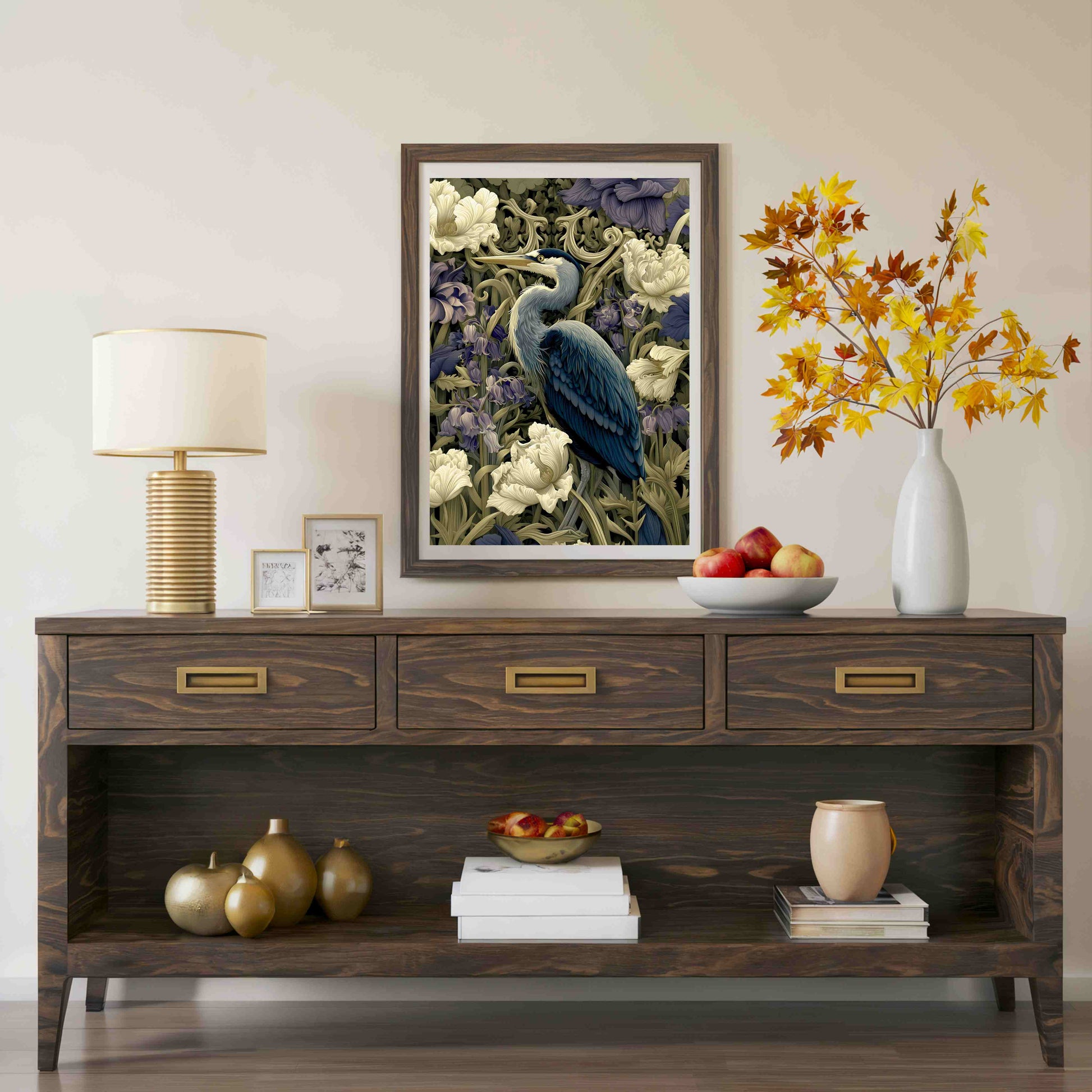 A framed art print depicting a blue heron among white flowers, styled on a dark wood console table in a living room setting with a lamp, decorative vases, and fruit.