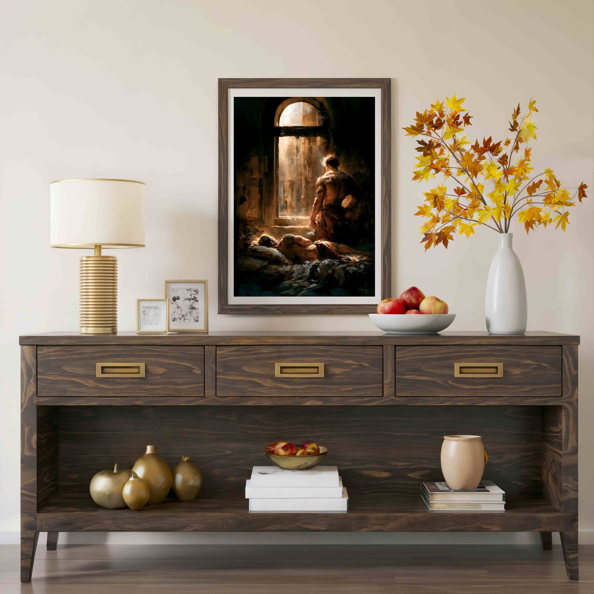 A framed art print depicting a biblical scene of a man in a dark cave, styled on a dark wood console table with a lamp, fruit, and decorative vases in a warm, neutral room setting.