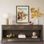 A dark brown wooden console table styled in a living room setting, featuring a vintage 'Vintage' magazine poster hanging above it, a table lamp, decorative vases, and fruit bowls.