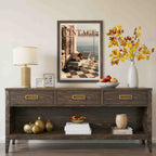 A dark brown wooden console table styled in a living room setting, featuring a matching framed art print on the wall above it. The table is decorated with a white table lamp, small picture frames, a white vase with autumn-colored leaves, a bowl of apples, and decorative books.