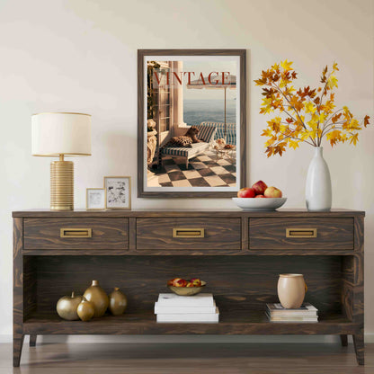A dark brown wooden console table styled in a living room setting, featuring a matching framed art print on the wall above it. The table is decorated with a white table lamp, small picture frames, a white vase with autumn-colored leaves, a bowl of apples, and decorative books.