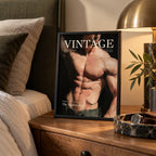 A framed art print featuring a muscular male torso with the text 'VINTAGE' and 'The Modern Man' is styled on a wooden nightstand next to a bed, a lamp, and a plant.