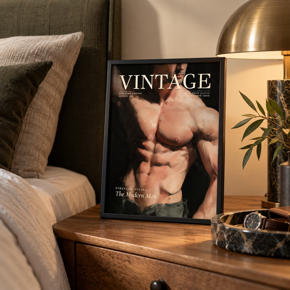 A framed art print featuring a muscular male torso with the text 'VINTAGE' and 'The Modern Man' is styled on a wooden nightstand next to a bed, a lamp, and a plant.