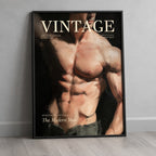 A framed vintage-style poster leaning against a light gray wall on a pale wood floor. The poster features a muscular male torso with the word 'VINTAGE' across the top and additional text including 'The Modern Man'.