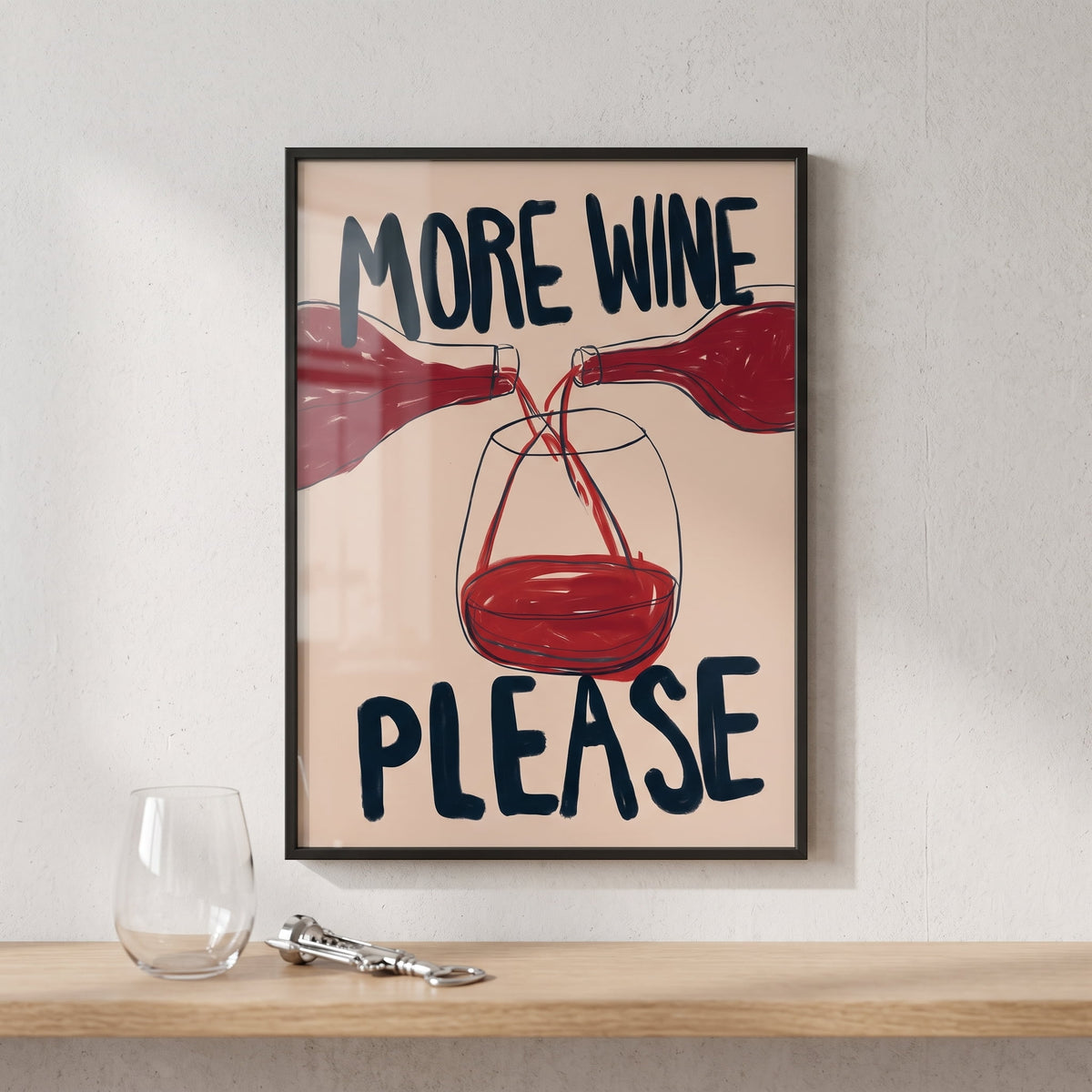 A framed art print with the text 'MORE WINE PLEASE' and an illustration of red wine being poured into a glass, displayed on a wooden shelf next to an empty wine glass and a corkscrew against a textured white wall.