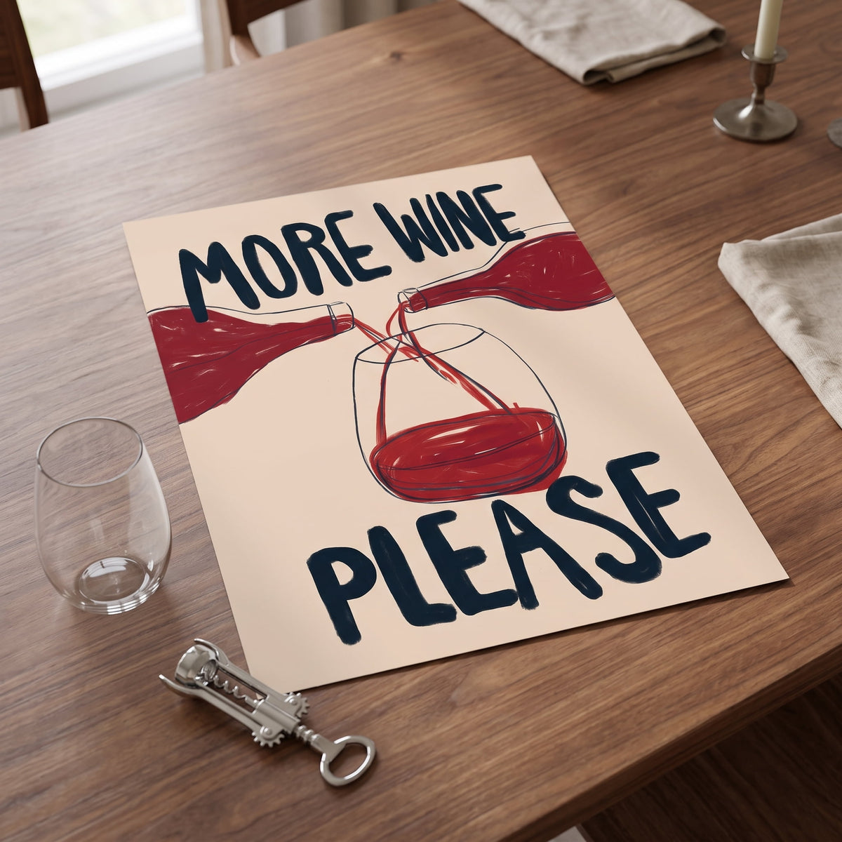 A rectangular printed sign or placemat on a wooden dining table, featuring the phrase “MORE WINE PLEASE” in dark blue lettering above and below an illustration of red wine bottles pouring into a wine glass. A clear wine glass, corkscrew, and candlestick are arranged around it in a real indoor setting.