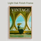 A framed vintage-style poster with the word VINTAGE at the top and a woman seated inside an arched window scene. The frame is labeled as light oak finish, and the poster is shown against a plain light gray background.