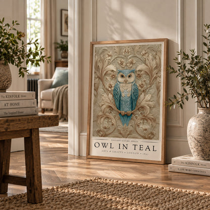 A framed art print featuring a blue owl on a branch with ornate floral details, leaning against a white wall in a styled room with wood floors, plants, and books.
