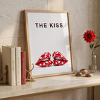 A framed art print leaning on a marble shelf against a beige tiled wall. The print features the text 'THE KISS' above a pair of red lips with white polka dots.