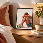 Small Rest gay art print in a black frame on a wooden bedside table under a warm lamp beside a plant and mug, a seated man resting in colourful trainers