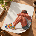Unframed Rest poster curling on a wooden desk beside coffee and a plant, an athletic man sitting with his head on his knees in multicoloured running trainers