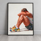 Rest gay art print in a black frame leaning against a plain grey wall on a pale wood floor, a seated man resting his head on his knees in colourful trainers