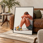 Rest gay art print in a black frame leaning against a vintage brown leather sofa in a warm living room, a seated man resting his head on his knees