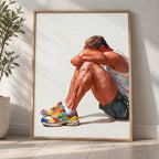 Rest gay art print in a light oak frame leaning against a white wall beside an olive tree, a seated man resting his head on his knees in colourful trainers