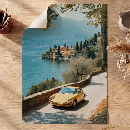 Vintage yellow porsche on a scenic road with a lake and buildings in the background, placed on a wooden surface with decorative items.