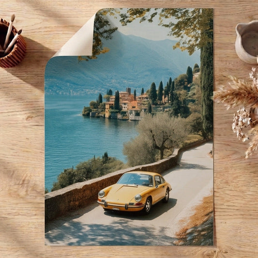 Vintage yellow porsche on a scenic road with a lake and buildings in the background, placed on a wooden surface with decorative items.