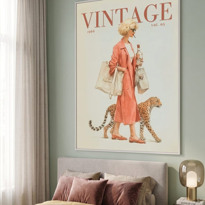 A framed vintage-style wall art print hangs above a bed in a styled bedroom. The print features the word VINTAGE at the top and an illustration of a woman in a peach dress walking with a leopard, holding shopping bags.