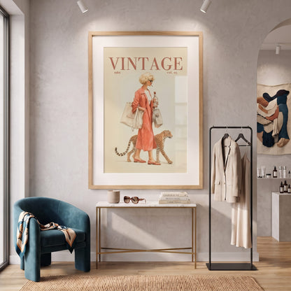 A large framed vintage-style wall art print hangs on a textured light gray wall in a styled interior. The artwork shows a woman in a pink dress walking with a cat, with the word 'VINTAGE' at the top.