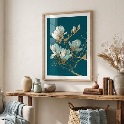 A framed art print featuring white magnolia blossoms on a dark teal background, styled on a light beige wall above a rustic wooden console table with decorative vases and dried grass.