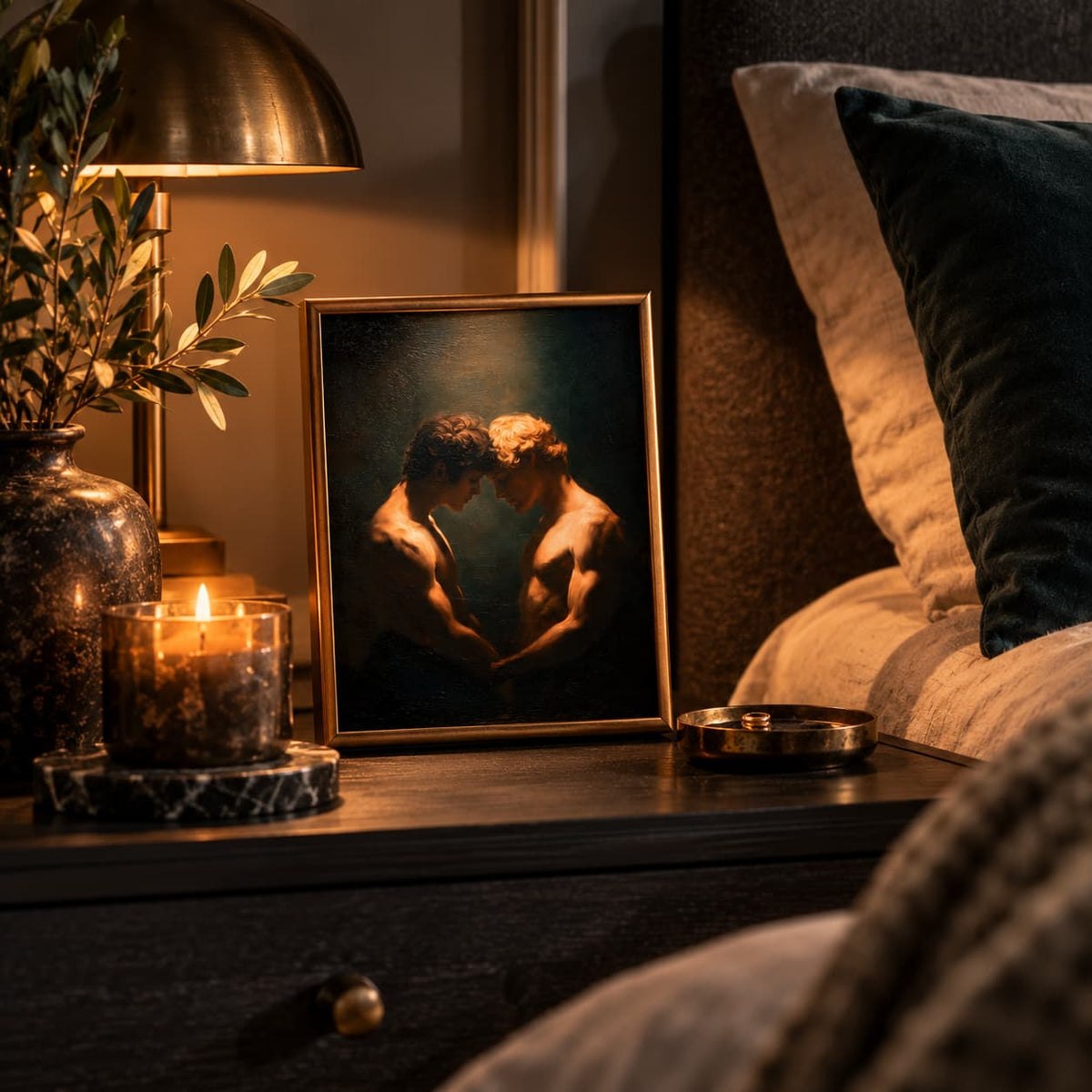 A framed art print depicting two shirtless men embracing in a dimly lit scene, styled on a dark wooden nightstand next to a lit candle, a brass table lamp, and a vase with greenery, with a bed visible in the background.
