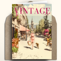 A vintage-style illustrated greeting card or magazine cover titled "VINTAGE" is shown standing in front of a brown envelope. The artwork depicts a sunlit street scene with a woman walking a dog, café seating, trees, and a church spire in the distance.