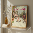 A framed vintage-style art print leaning against a wall on a glass table, next to a brown glass vase with dried branches and stacked books. The print features a retro street scene with the word 'VINTAGE' in pink at the top.