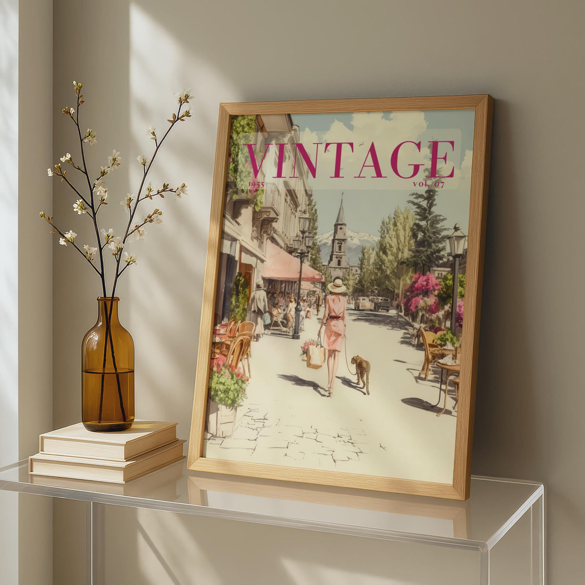 A framed vintage-style art print leaning against a wall on a glass table, next to a brown glass vase with dried branches and stacked books. The print features a retro street scene with the word 'VINTAGE' in pink at the top.