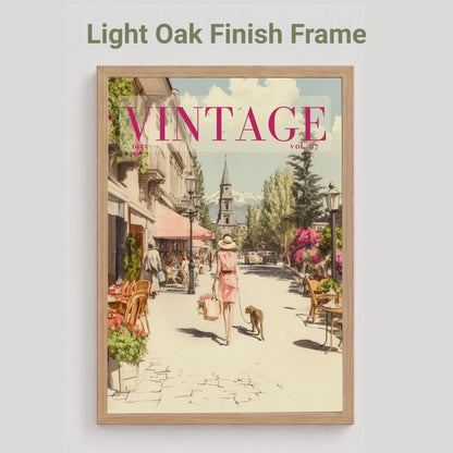 A framed art print featuring a vintage-style illustration of a woman walking down a cobblestone street with dogs and cafe tables. The print is housed in a light oak wood frame, shown against a neutral light gray background with the text 'Light Oak Finish Frame' above it.