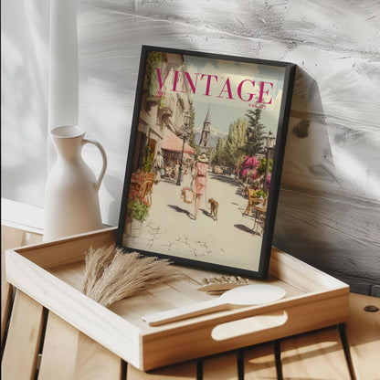 A framed art print featuring a vintage street scene with the word 'VINTAGE' in pink at the top, styled on a wooden tray next to a white ceramic pitcher against a textured gray wall.