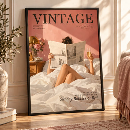 A framed vintage-style wall poster leaning against a wall in a sunlit bedroom. The poster shows a woman reclining in bed reading a newspaper and holding a glass, with a pink background and the word VINTAGE across the top.
