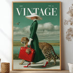 A framed vintage-style poster leaning against a wall on a shelf, surrounded by decorative dried flowers and small vases. The poster shows a woman in a long green dress and white hat walking beside a leopard while carrying a red suitcase, with the word VINTAGE at the top.
