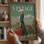 A person holds a framed art print featuring a vintage-style illustration with the word 'VINTAGE' at the top. The artwork shows a woman in a green dress and white hat carrying a red suitcase, accompanied by a leopard, against a teal sky and pale ground.
