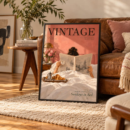 A framed vintage-style wall art print leaning against a brown leather sofa in a warm living room. The artwork shows a person reading a newspaper in bed with the word VINTAGE prominently displayed at the top.
