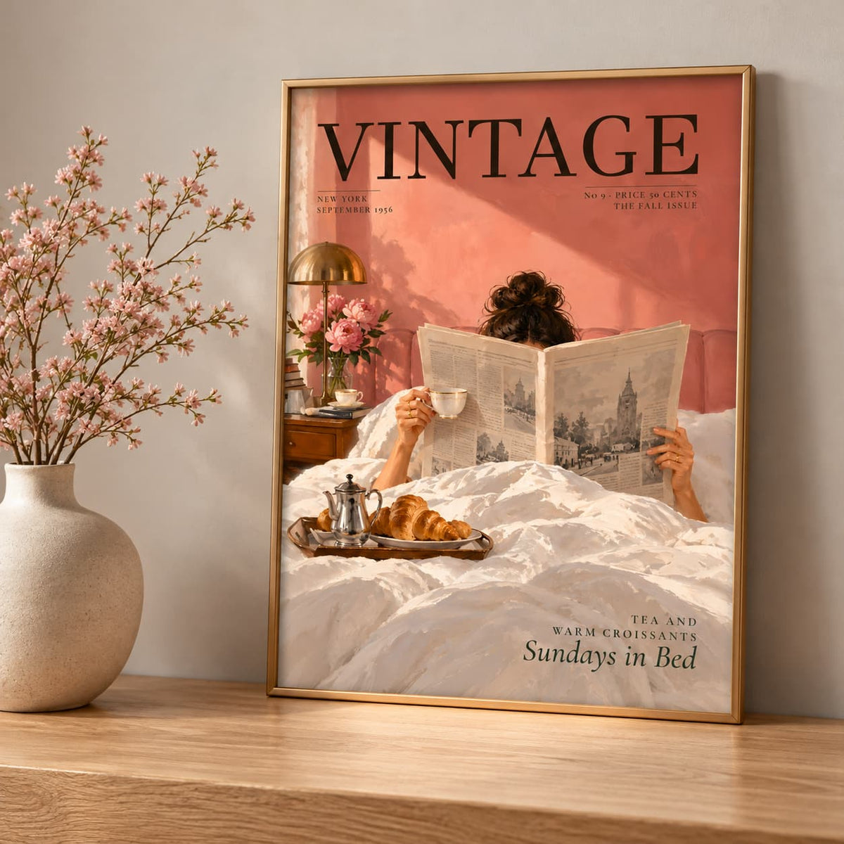 A framed vintage-style poster leaning against a wall on a wooden surface, next to a ceramic vase with pink blossoms. The poster shows a person reading a newspaper in bed with the title 'VINTAGE' at the top and the text 'Tea and Warm Croissants Sundays in Bed' at the bottom.