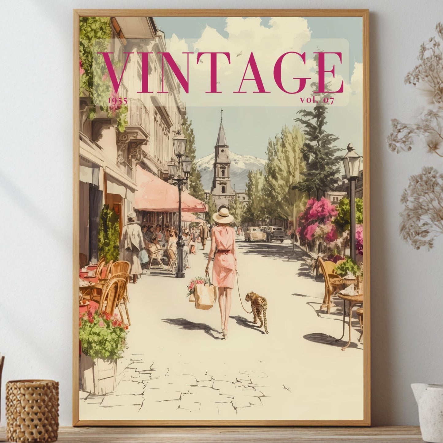 A framed vintage-style poster print leaning against a wall on a shelf, surrounded by decorative dried flowers and small vases. The poster shows a retro street scene with the word "VINTAGE" in large pink letters at the top.