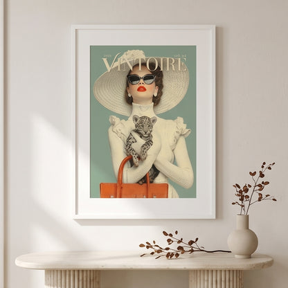A framed art print featuring a vintage Vogue cover illustration of a woman in a large sunhat and sunglasses holding a cat, displayed on a light wall above a console table with a vase and dried branches.