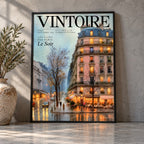 Black-framed Vintoire Rainy Paris café poster leaning against a grey plaster wall in soft window light, next to an olive tree in a stone urn.