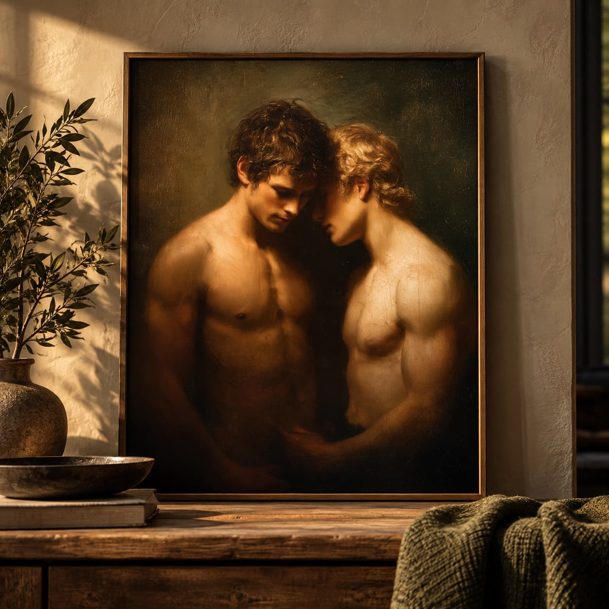 A framed classical-style painting depicting two shirtless men embracing, displayed on a wooden table in a warmly lit room with a plant and textured throw blanket nearby.