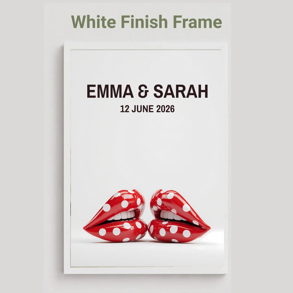 A framed custom wedding print with a white finish frame, featuring the text 'EMMA & SARAH' and '12 JUNE 2026' above a graphic of two red lips with white polka dots, on a light gray background.