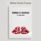 A framed custom wedding print with a white finish frame, featuring the text 'EMMA & SARAH' and '12 JUNE 2026' above a graphic of two red lips with white polka dots, on a light gray background.