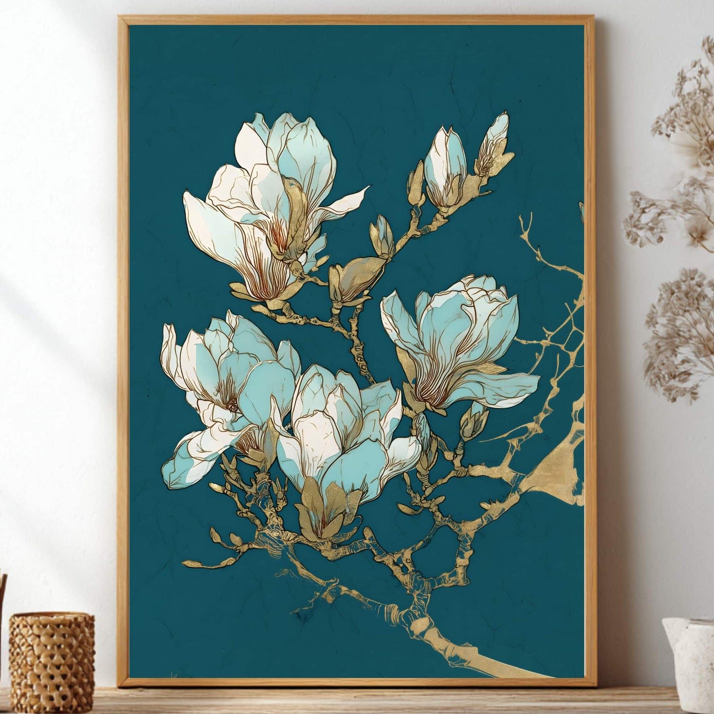 A framed botanical art print featuring light blue and white magnolia blossoms with gold accents on a deep teal background, styled on a shelf with dried flowers and small decorative vases.