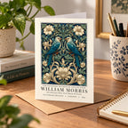 A William Morris art print card featuring two blue birds and white flowers on a dark blue background, displayed on a wooden desk with a plant, books, and stationery.