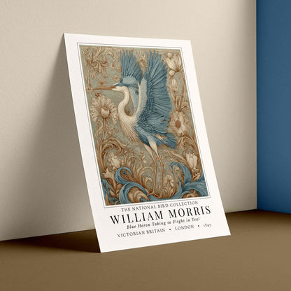 Decorative poster with a bird design leaning against a wall. William Morris style exhibition poster featuring a blue heron taking to flight, surrounded by ornate acanthus scrolls, dahlias and botanical foliage in teal, gold and antique cream — Arts & Crafts wall art print