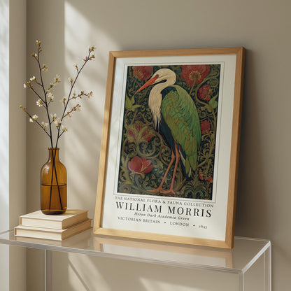 Framed artwork of a heron by William Morris on a shelf with a vase and books.William Morris inspired heron exhibition poster in a jewel-toned palette of forest green, antique crimson and old gold, featuring a grey heron amid ornate pimpernel blooms and curling vines — Victorian Arts and Crafts style, framed print.