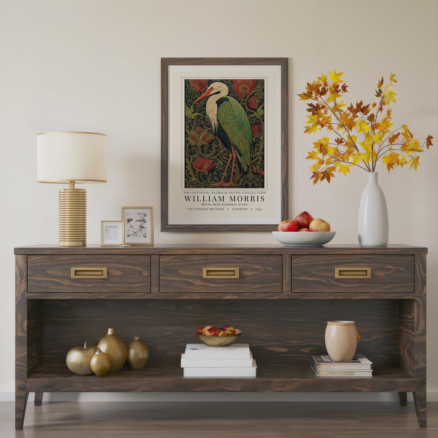 Wooden console table with decorative items including a lamp, framed artwork, and a vase with autumn leaves. William Morris inspired heron exhibition poster in a jewel-toned palette of forest green, antique crimson and old gold, featuring a grey heron amid ornate pimpernel blooms and curling vines — Victorian Arts and Crafts style, framed print.